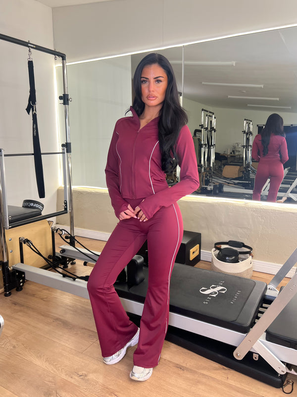 Woman in a maroon tracksuit standing in a Pilates reformer room.