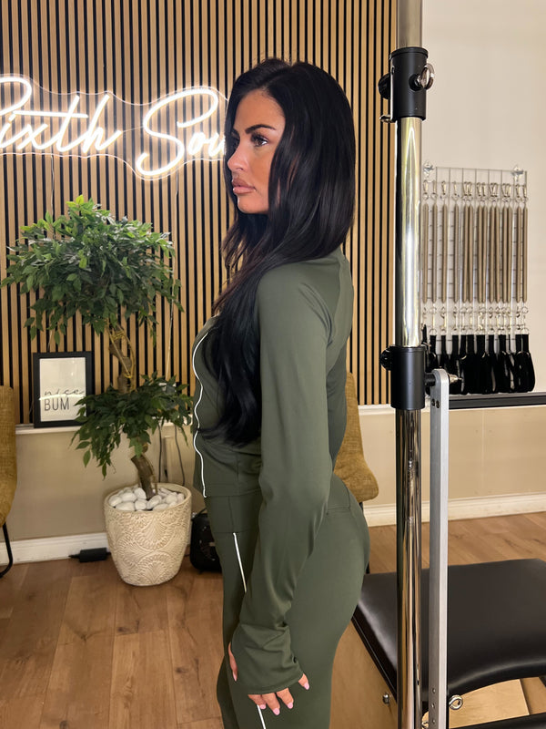 Woman in a green outfit standing in a room with plants and a neon sign.