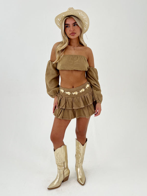 Woman wearing a camel outfit with a matching hat gold belt and boots on a white background