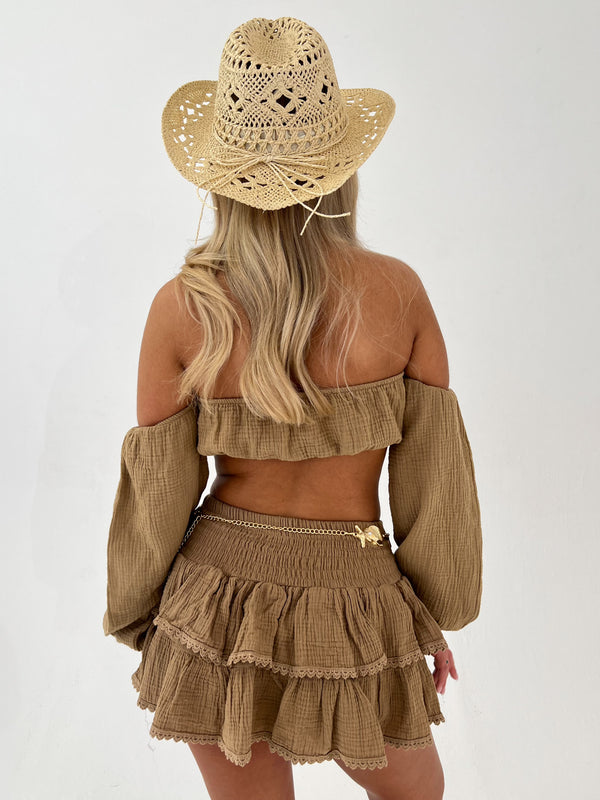 Woman wearing a camel off-shoulder top and skirt with a straw hat on a white background