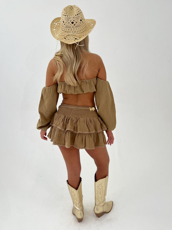 Woman wearing a camel outfit with a matching hat gold belt and boots on a white background