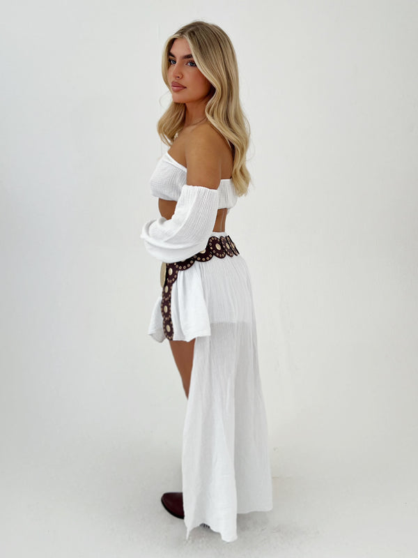 Woman wearing a white off-shoulder top and sheer white skirt with a patterned belt on a plain background
