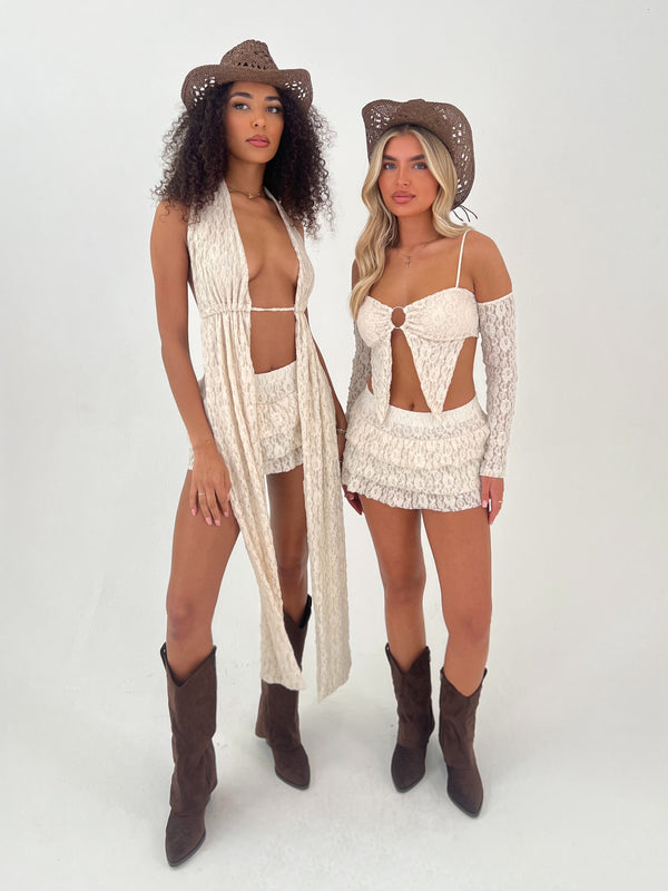 Two women wearing matching beige lace outfits with cowboy hats and boots on a white background