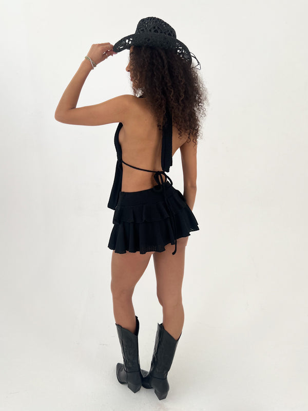 Woman wearing a black outfit with ruffles and black boots, holding a black hat against a white background