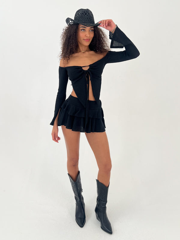 Woman wearing a black outfit with a skirt and long-sleeve top, holding a black hat on a white background.